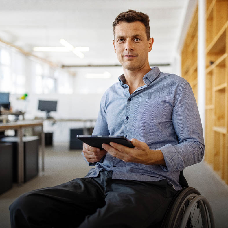 In a comfortable office space, a man on a wheelchair is learning about the service options for the mobility impaired and smiles holding his tablet.